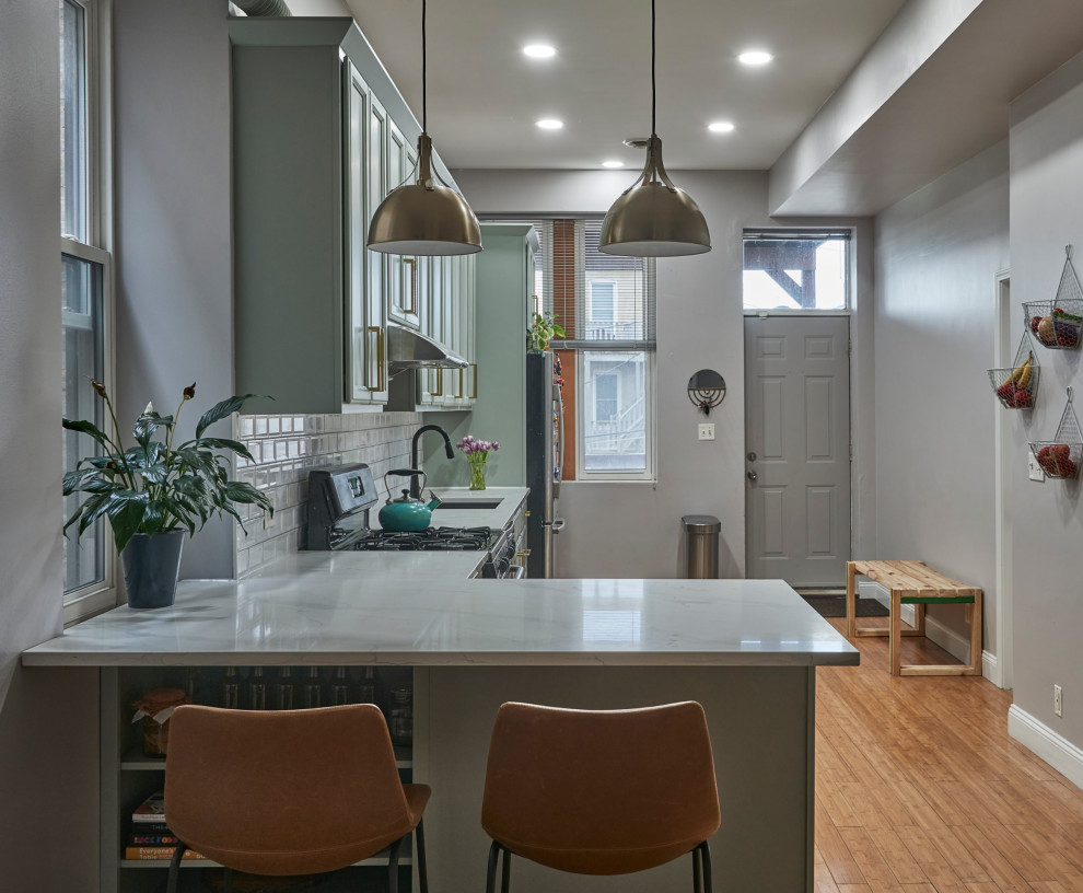 This is an example of a mid-sized transitional l-shaped open plan kitchen in Chicago with an undermount sink, recessed-panel cabinets, green cabinets, quartzite benchtops, white splashback, subway tile splashback, black appliances, medium hardwood floors, a peninsula and white benchtop.
