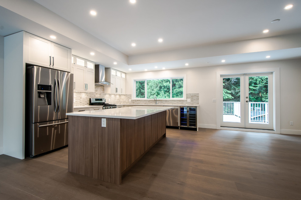 Large contemporary l-shaped open plan kitchen in Vancouver with an undermount sink, shaker cabinets, white cabinets, stainless steel appliances, with island, grey splashback, matchstick tile splashback, medium hardwood floors and quartz benchtops.