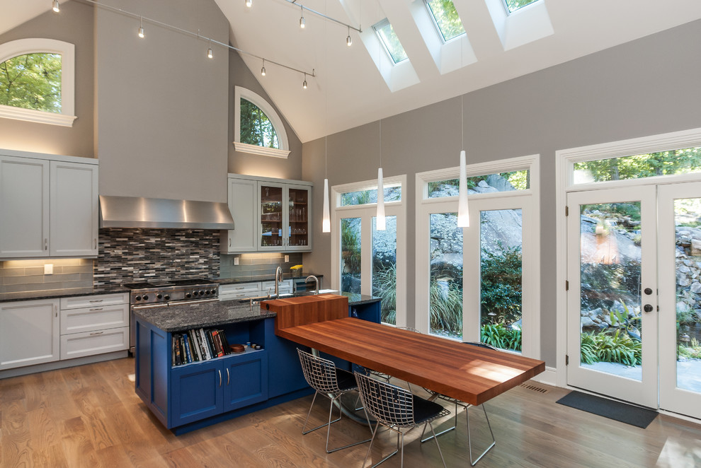 Large danish l-shaped medium tone wood floor open concept kitchen photo in Raleigh with an undermount sink, recessed-panel cabinets, blue cabinets, wood countertops, blue backsplash, glass tile backsplash, stainless steel appliances and an island
