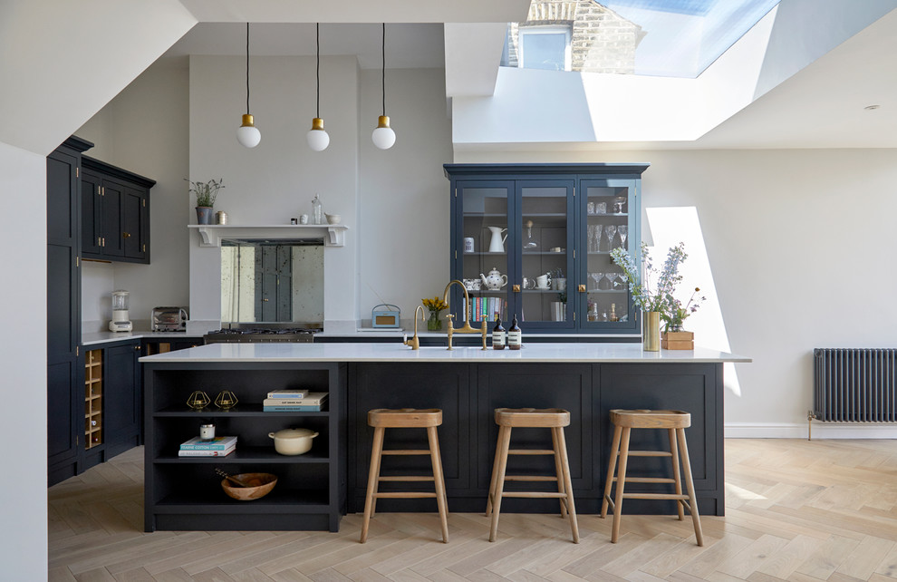 Transitional l-shaped kitchen in London with shaker cabinets, blue cabinets, metallic splashback, mirror splashback, light hardwood floors, with island, beige floor and white benchtop.