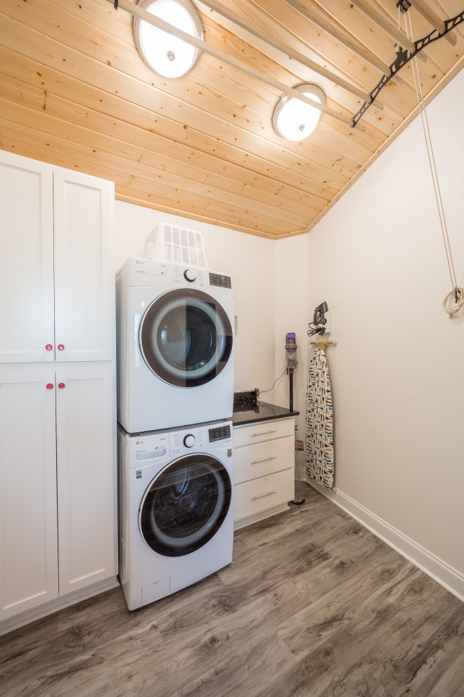 This is an example of a mid-sized traditional single-wall dedicated laundry room with flat-panel cabinets, white cabinets, granite benchtops, white walls, vinyl floors, a stacked washer and dryer, grey floor, black benchtop and wood.