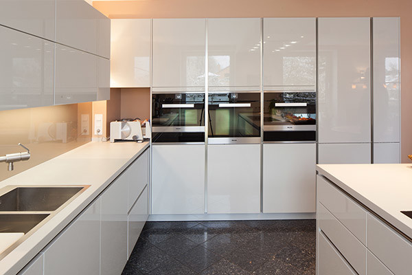 Küche in L-Form mit integriertem Waschbecken, flächenbündigen Schrankfronten, weißen Schränken, Quarzwerkstein-Arbeitsplatte, Küchenrückwand in Beige, Küchengeräten aus Edelstahl, Marmorboden, Kücheninsel, Glasrückwand und schwarzem Boden