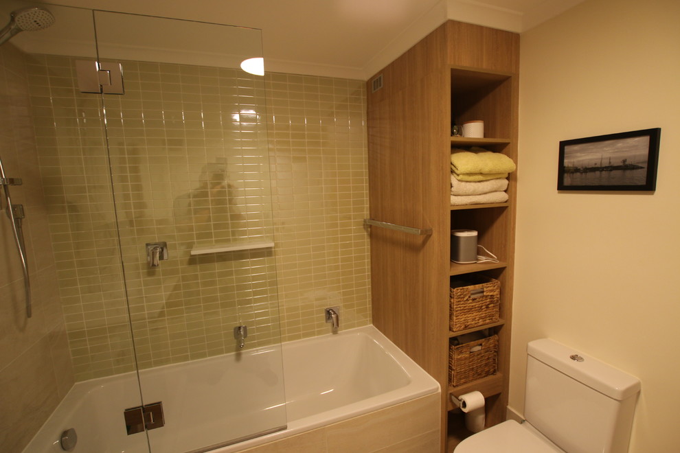 Mid-sized contemporary master bathroom in Sydney with flat-panel cabinets, medium wood cabinets, a corner tub, a shower/bathtub combo, a one-piece toilet, green tile, ceramic tile, beige walls, ceramic floors, an integrated sink, solid surface benchtops, beige floor, a hinged shower door and white benchtops.