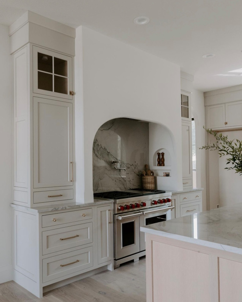 Inredning av ett modernt stort vit vitt kök, med en undermonterad diskho, luckor med infälld panel, vita skåp, marmorbänkskiva, flerfärgad stänkskydd, stänkskydd i marmor, rostfria vitvaror, ljust trägolv, en köksö och flerfärgat golv