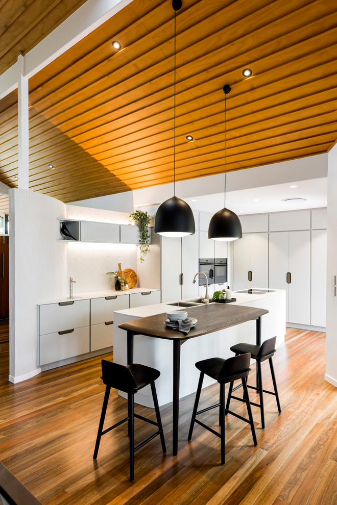 Photo of a mid-sized contemporary l-shaped eat-in kitchen in Brisbane with an undermount sink, flat-panel cabinets, white cabinets, white splashback, black appliances, medium hardwood floors, with island, brown floor and white benchtop.