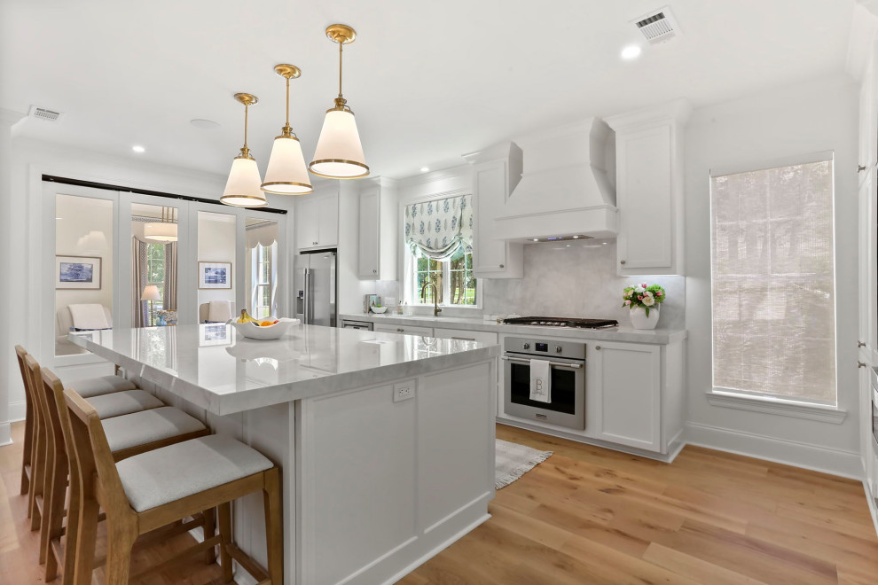 Large transitional l-shaped light wood floor and brown floor open concept kitchen photo in New Orleans with an undermount sink, shaker cabinets, white cabinets, quartz countertops, white backsplash, quartz backsplash, stainless steel appliances, an island and white countertops