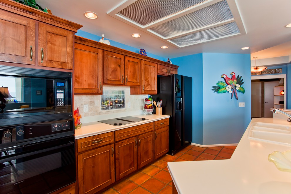 Photo of a mid-sized tropical galley open plan kitchen in San Diego with a triple-bowl sink, shaker cabinets, medium wood cabinets, solid surface benchtops, beige splashback, stone tile splashback, black appliances and terra-cotta floors.