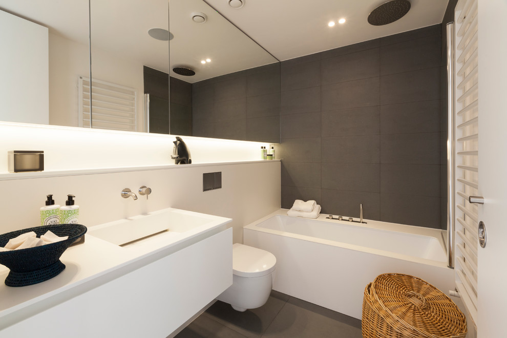 This is an example of a mid-sized contemporary kids bathroom in London with a wall-mount sink, furniture-like cabinets, white cabinets, solid surface benchtops, a corner tub, a wall-mount toilet, gray tile, stone tile, white walls and slate floors.