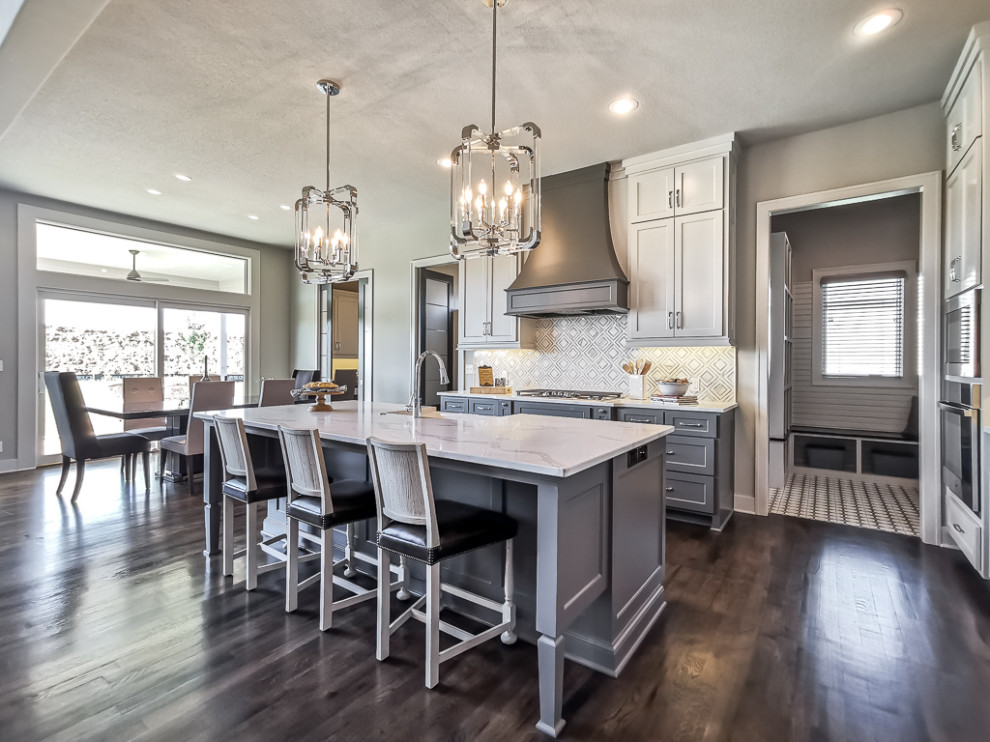 This is an example of a transitional l-shaped kitchen in Kansas City with a farmhouse sink, shaker cabinets, white cabinets, grey splashback, stainless steel appliances, dark hardwood floors, with island, brown floor and white benchtop.