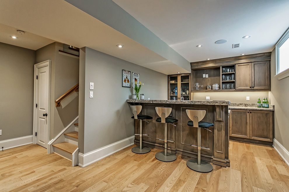 This is an example of a mid-sized transitional galley seated home bar in Toronto with shaker cabinets, medium wood cabinets, light hardwood floors, granite benchtops and beige floor.