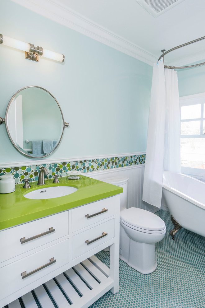 Photo of a small transitional kids bathroom in Los Angeles with furniture-like cabinets, medium wood cabinets, a hot tub, a shower/bathtub combo, a one-piece toilet, green tile, glass sheet wall, blue walls, mosaic tile floors, an undermount sink, engineered quartz benchtops and blue floor.