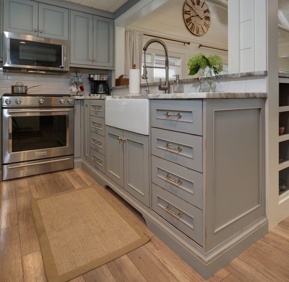 This is an example of a small beach style u-shaped eat-in kitchen in Detroit with a farmhouse sink, beaded inset cabinets, distressed cabinets, granite benchtops, white splashback, subway tile splashback, stainless steel appliances, light hardwood floors, no island, brown floor and grey benchtop.