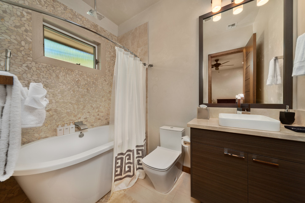 Mid-sized contemporary 3/4 bathroom in Hawaii with porcelain floors, flat-panel cabinets, brown cabinets, a freestanding tub, a shower/bathtub combo, a one-piece toilet, beige tile, pebble tile, beige walls, a vessel sink, limestone benchtops, beige floor and a shower curtain.