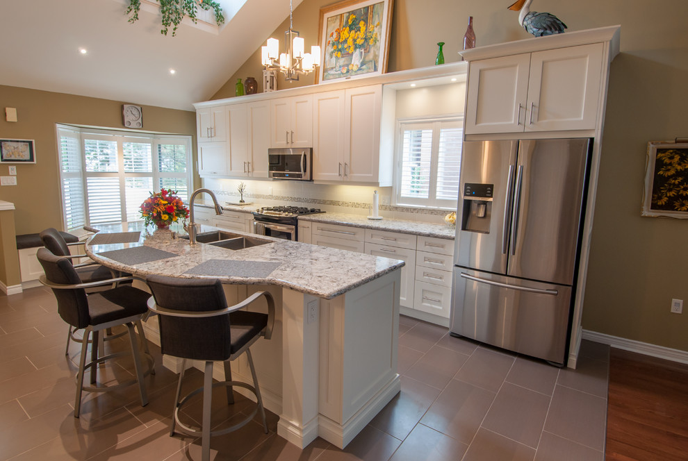 Inspiration for a mid-sized transitional galley ceramic tile and gray floor eat-in kitchen remodel in Toronto with a double-bowl sink, shaker cabinets, white cabinets, beige backsplash, stone tile backsplash, stainless steel appliances, an island and granite countertops