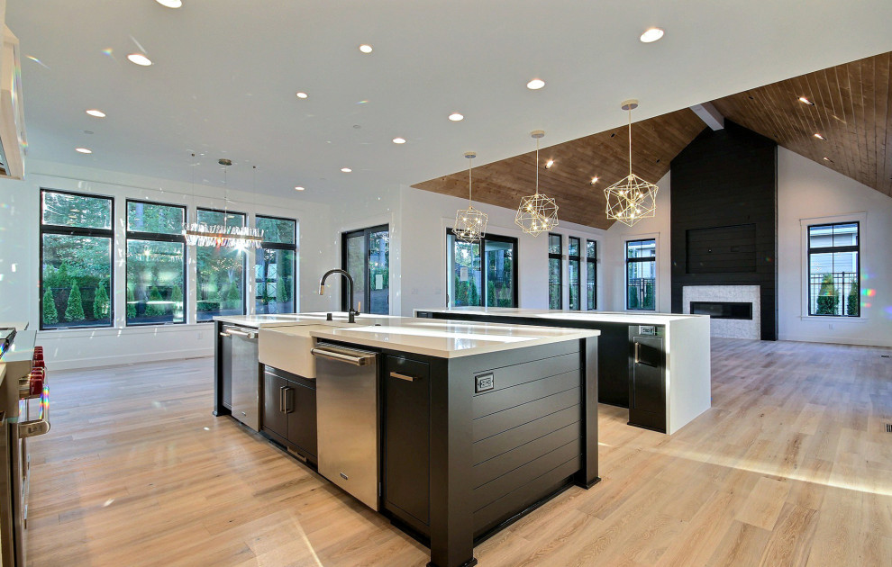 This is an example of an expansive country single-wall eat-in kitchen in Portland with a farmhouse sink, flat-panel cabinets, black cabinets, quartz benchtops, white splashback, ceramic splashback, stainless steel appliances, light hardwood floors, multiple islands, beige floor and white benchtop.