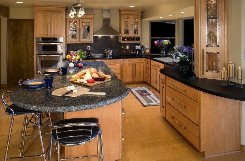 Example of a mid-sized l-shaped medium tone wood floor kitchen design with flat-panel cabinets, medium tone wood cabinets, black backsplash, stone tile backsplash, stainless steel appliances and an island