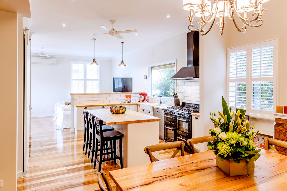 This is an example of a contemporary l-shaped eat-in kitchen in Melbourne with a farmhouse sink, white cabinets, wood benchtops, white splashback, subway tile splashback, black appliances, medium hardwood floors, with island and recessed-panel cabinets.