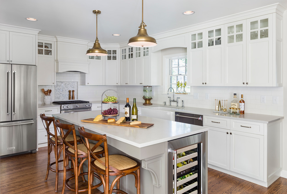 Photo of a transitional l-shaped kitchen in Minneapolis with a farmhouse sink, recessed-panel cabinets, white cabinets, white splashback, subway tile splashback, stainless steel appliances, dark hardwood floors, with island, brown floor and grey benchtop.