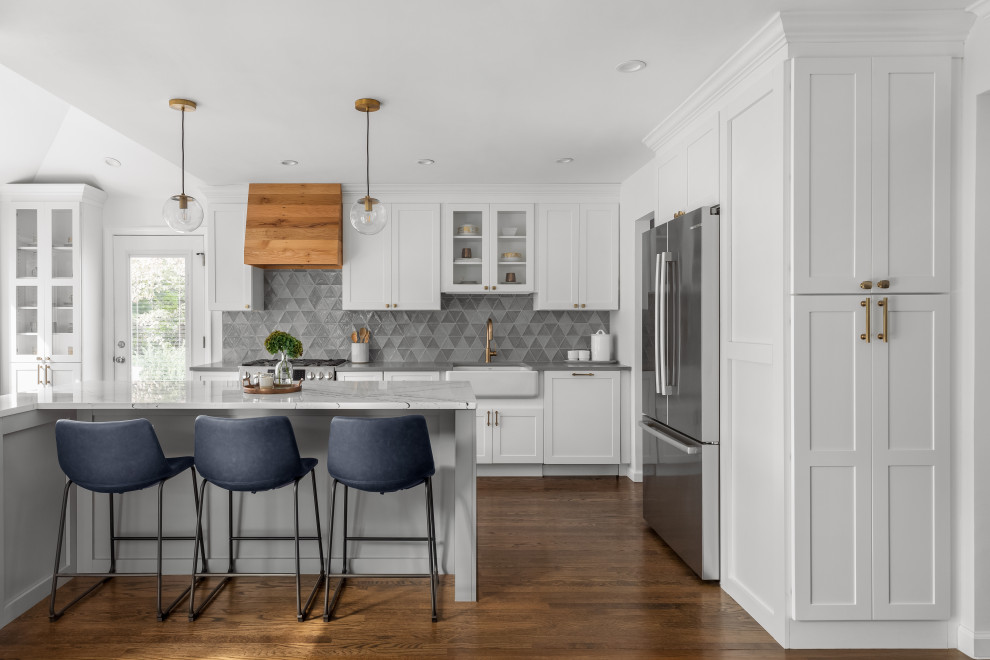 Inspiration for a mid-sized transitional l-shaped open plan kitchen in New York with a farmhouse sink, shaker cabinets, white cabinets, quartz benchtops, grey splashback, glass tile splashback, stainless steel appliances, dark hardwood floors, with island, brown floor and grey benchtop.