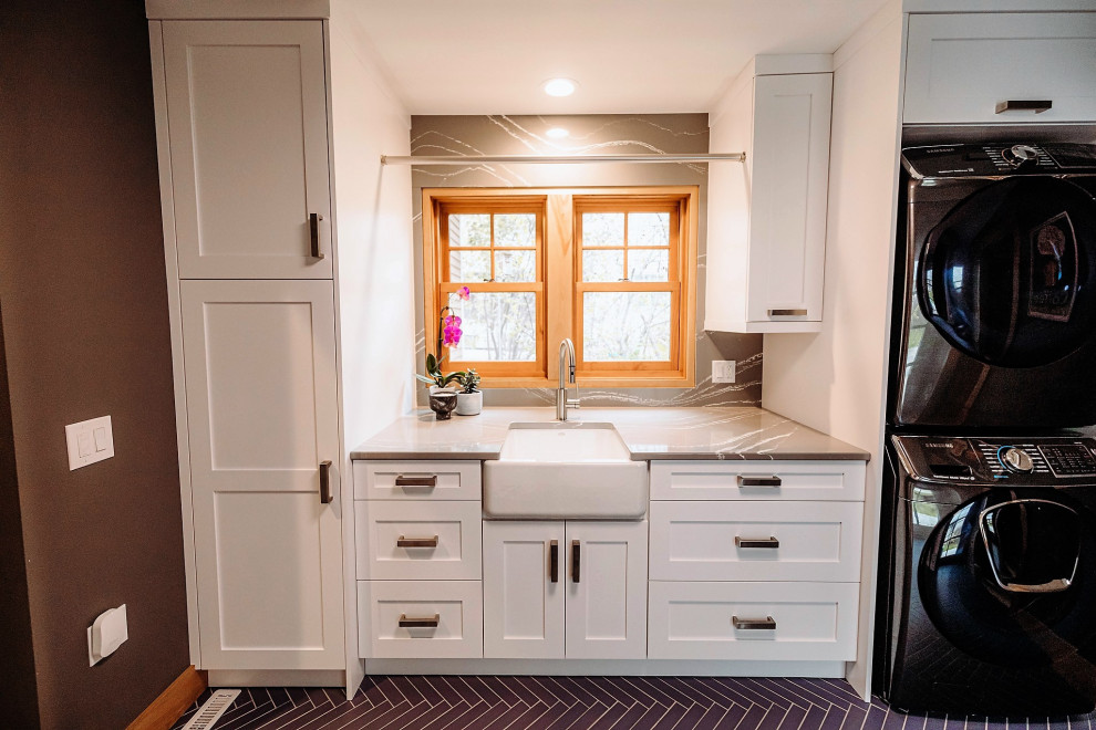 Example of a large arts and crafts galley porcelain tile and blue floor utility room design in Other with a farmhouse sink, shaker cabinets, white cabinets, quartz countertops, gray backsplash, quartz backsplash, gray walls, a stacked washer/dryer and gray countertops