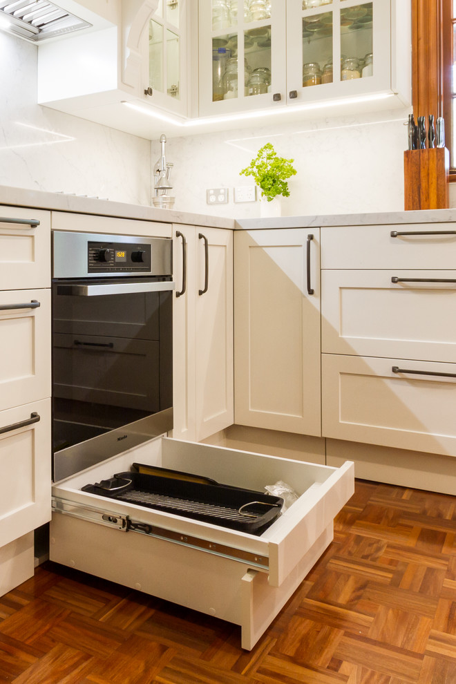 This is an example of a mid-sized traditional u-shaped eat-in kitchen in Melbourne with a drop-in sink, shaker cabinets, white cabinets, marble benchtops, white splashback, marble splashback, stainless steel appliances, medium hardwood floors, a peninsula, brown floor and white benchtop.