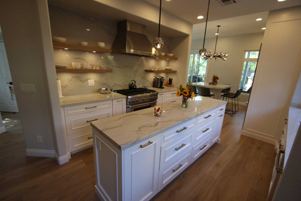 Inspiration for a mid-sized transitional galley eat-in kitchen in Orange County with a farmhouse sink, shaker cabinets, white cabinets, granite benchtops, white splashback, granite splashback, stainless steel appliances, light hardwood floors, multiple islands, brown floor, white benchtop and wood.