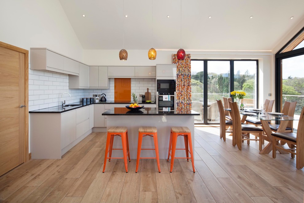 Mid-sized contemporary l-shaped open plan kitchen in Cornwall with flat-panel cabinets, grey cabinets, onyx benchtops, orange splashback, travertine splashback, black appliances, with island, black benchtop, an undermount sink, light hardwood floors and beige floor.