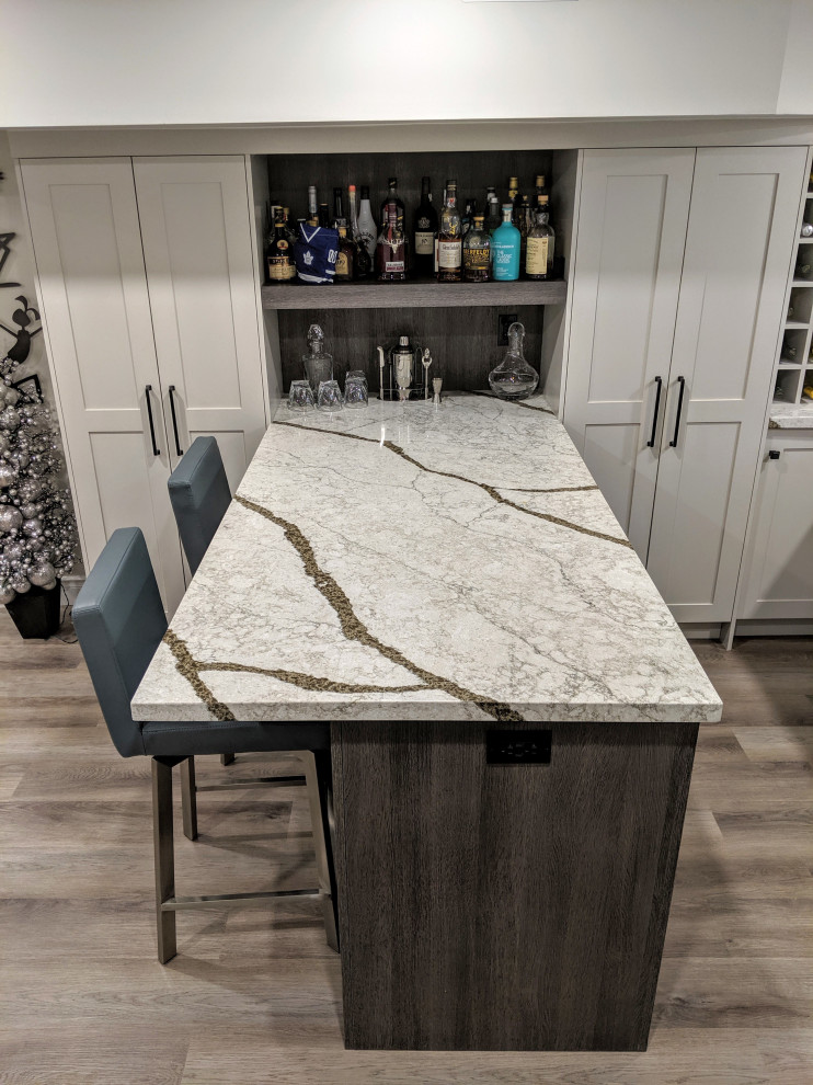 This is an example of a mid-sized transitional l-shaped wet bar in Toronto with an undermount sink, recessed-panel cabinets, grey cabinets, quartz benchtops, multi-coloured splashback, cement tile splashback, vinyl floors, brown floor and white benchtop.