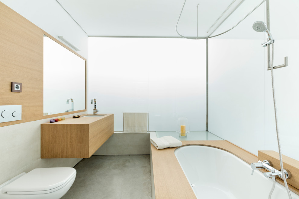 This is an example of a mid-sized contemporary bathroom in London with a curbless shower, a wall-mount toilet, white tile, glass sheet wall, white walls, concrete floors, a drop-in sink, wood benchtops, an undermount tub and brown benchtops.