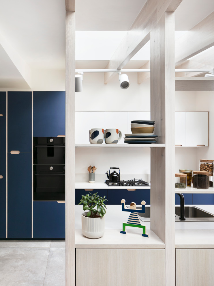 Design ideas for a mid-sized scandinavian galley open plan kitchen in London with a drop-in sink, flat-panel cabinets, blue cabinets, solid surface benchtops, white splashback, granite splashback, black appliances, porcelain floors, no island, grey floor, white benchtop and exposed beam.