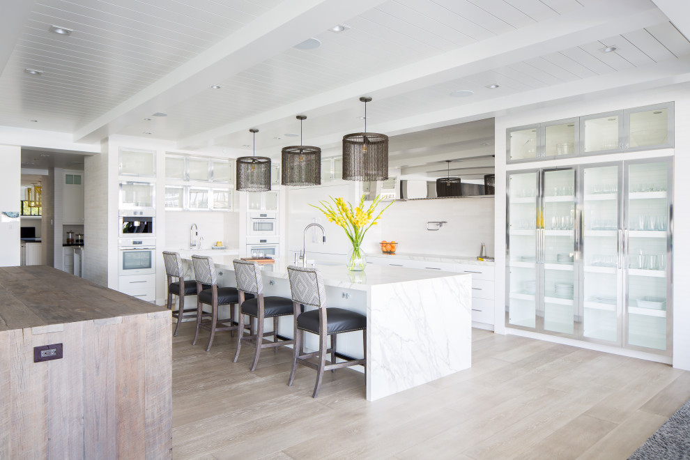 Photo of an expansive beach style l-shaped open plan kitchen in Orange County with glass-front cabinets, white cabinets, marble benchtops, white appliances, multiple islands and white benchtop.