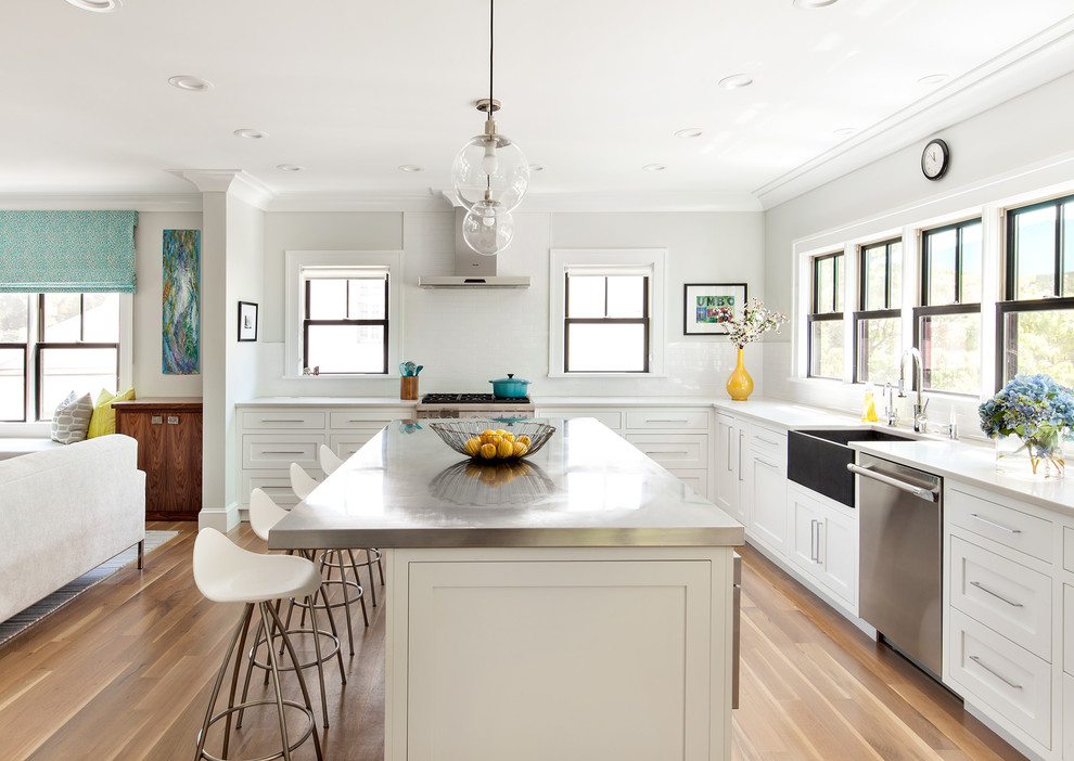 This is an example of a transitional l-shaped open plan kitchen in New York with a farmhouse sink, shaker cabinets, white cabinets, window splashback, stainless steel appliances, light hardwood floors, with island, beige floor and stainless steel benchtops.