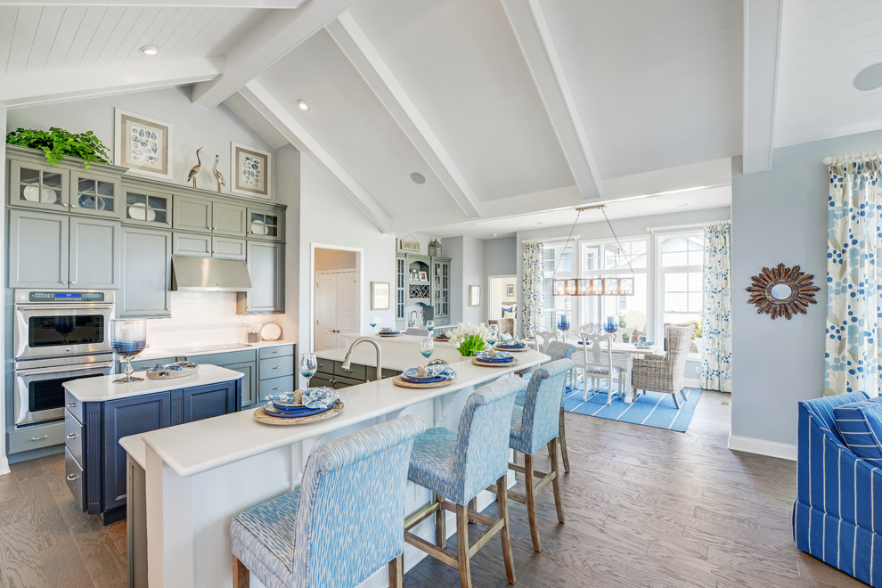 Photo of a beach style eat-in kitchen in Other with a farmhouse sink, recessed-panel cabinets, grey cabinets, quartz benchtops, white splashback, ceramic splashback, stainless steel appliances, medium hardwood floors and with island.