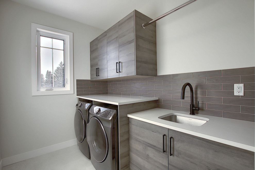 Mid-sized industrial single-wall dedicated laundry room in Calgary with an undermount sink, flat-panel cabinets, medium wood cabinets, quartz benchtops, grey splashback, cement tile splashback, grey walls, ceramic floors, a side-by-side washer and dryer, grey floor and white benchtop.