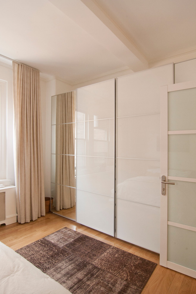This is an example of a mid-sized transitional gender-neutral built-in wardrobe in New York with glass-front cabinets, white cabinets, light hardwood floors and beige floor.