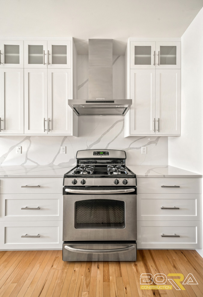 Example of a mid-sized single-wall light wood floor and brown floor eat-in kitchen design in Seattle with a farmhouse sink, shaker cabinets, white cabinets, quartz countertops, white backsplash, quartz backsplash, stainless steel appliances, an island and white countertops