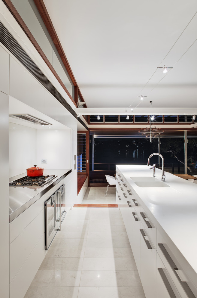 Photo of an expansive modern single-wall open plan kitchen in Sydney with an integrated sink, flat-panel cabinets, white cabinets, solid surface benchtops, white splashback, glass sheet splashback, stainless steel appliances, travertine floors and with island.