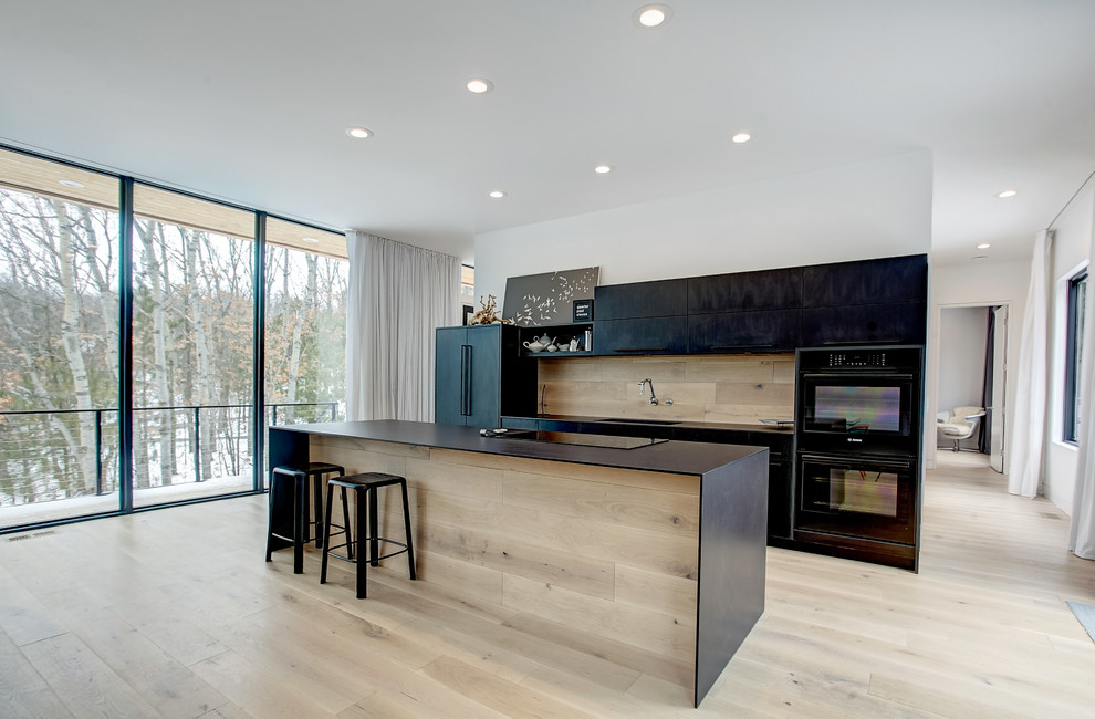 Inspiration for a mid-sized modern galley open plan kitchen in Grand Rapids with an integrated sink, flat-panel cabinets, black cabinets, quartz benchtops, timber splashback, black appliances, light hardwood floors, with island and beige floor.