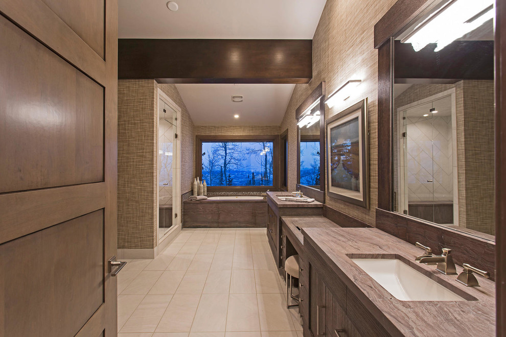 Expansive contemporary master bathroom in Salt Lake City with flat-panel cabinets, medium wood cabinets, a drop-in tub, a corner shower, a one-piece toilet, gray tile, ceramic tile, brown walls, ceramic floors, a drop-in sink, granite benchtops, beige floor, a hinged shower door, brown benchtops, a shower seat, a double vanity, a built-in vanity, exposed beam and wallpaper.