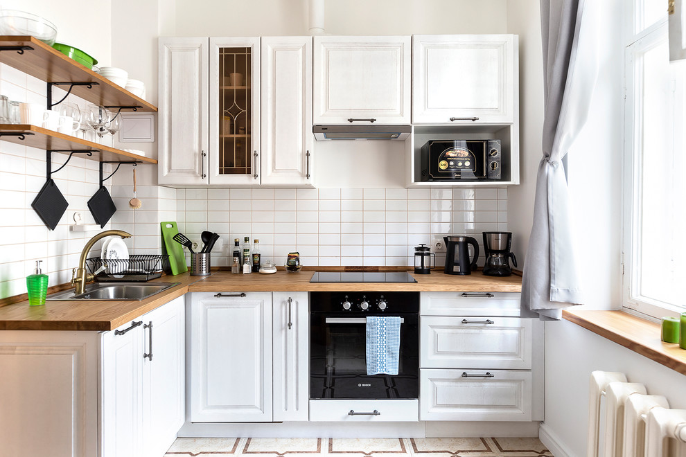 This is an example of a mid-sized scandinavian l-shaped eat-in kitchen in Saint Petersburg with a drop-in sink, raised-panel cabinets, white cabinets, wood benchtops, white splashback, subway tile splashback, black appliances, ceramic floors, no island, beige floor and brown benchtop.
