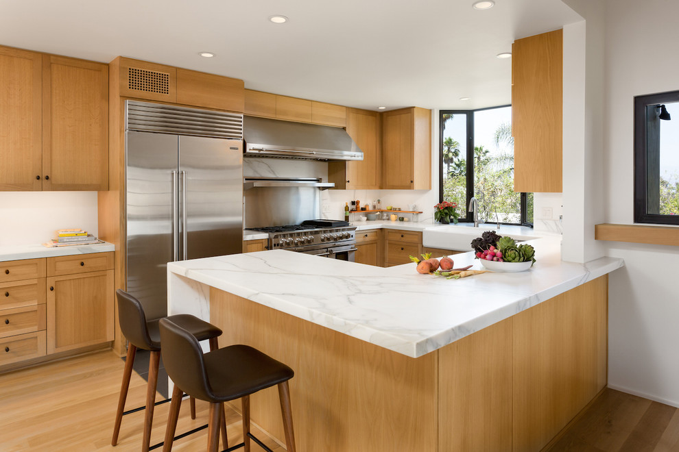 Inspiration for a mid-sized midcentury kitchen in Los Angeles with a farmhouse sink, shaker cabinets, light wood cabinets, marble benchtops, stainless steel appliances, light hardwood floors, a peninsula and metallic splashback.