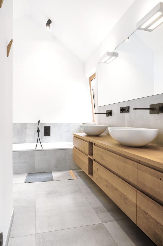 Пример оригинального дизайна: ванная комната среднего размера в стиле модернизм с плоскими фасадами, светлыми деревянными фасадами, накладной ванной, душем без бортиков, инсталляцией, серой плиткой, цементной плиткой, серыми стенами, полом из цементной плитки, настольной раковиной, столешницей из дерева, серым полом, открытым душем, тумбой под две раковины и подвесной тумбой