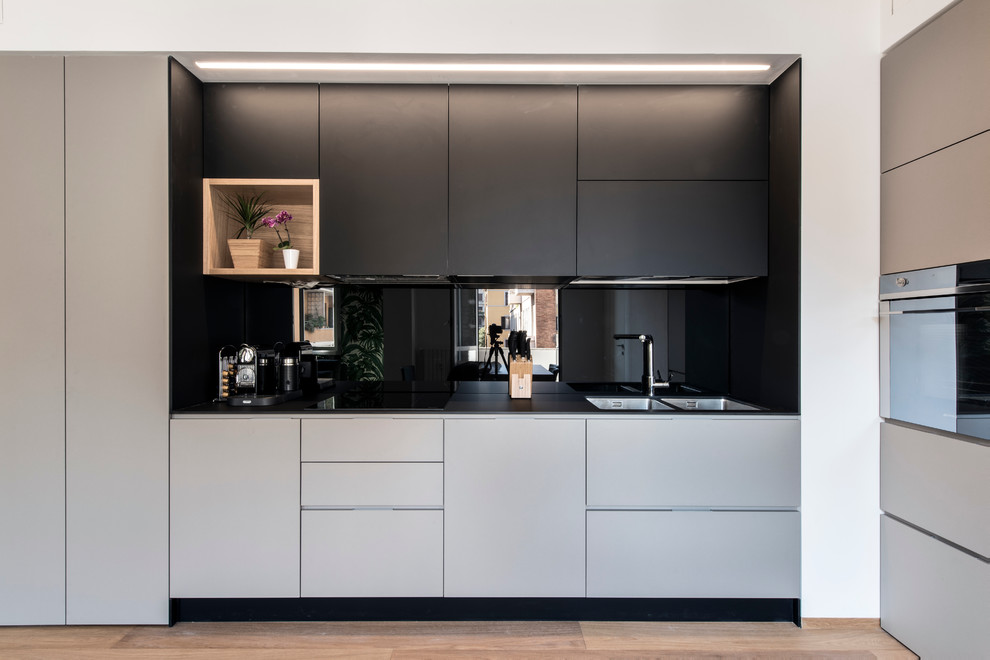 This is an example of a mid-sized contemporary l-shaped open plan kitchen in Milan with a double-bowl sink, flat-panel cabinets, black cabinets, laminate benchtops, black splashback, glass sheet splashback, stainless steel appliances, light hardwood floors and no island.