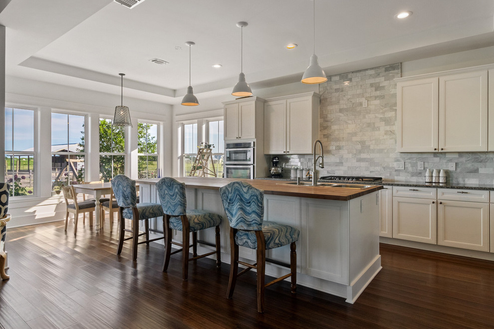 Inspiration for a mid-sized transitional l-shaped open plan kitchen in Austin with a triple-bowl sink, recessed-panel cabinets, white cabinets, granite benchtops, grey splashback, stone tile splashback, stainless steel appliances, dark hardwood floors, with island, brown floor and grey benchtop.