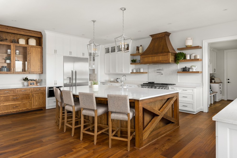 Design ideas for a country l-shaped kitchen in San Francisco with a farmhouse sink, shaker cabinets, white cabinets, white splashback, stainless steel appliances, medium hardwood floors, with island, brown floor and white benchtop.