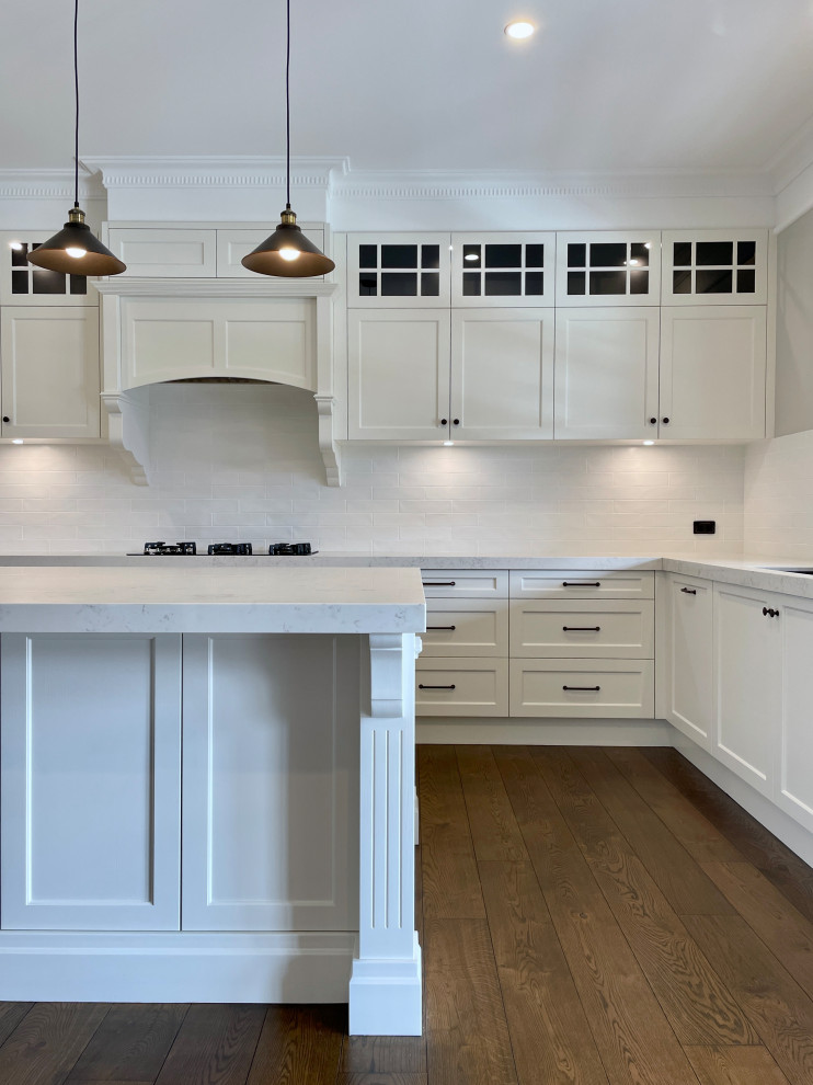 This is an example of an expansive traditional u-shaped open plan kitchen in Sydney with an undermount sink, recessed-panel cabinets, white cabinets, quartz benchtops, white splashback, subway tile splashback, black appliances, medium hardwood floors, with island, brown floor and white benchtop.