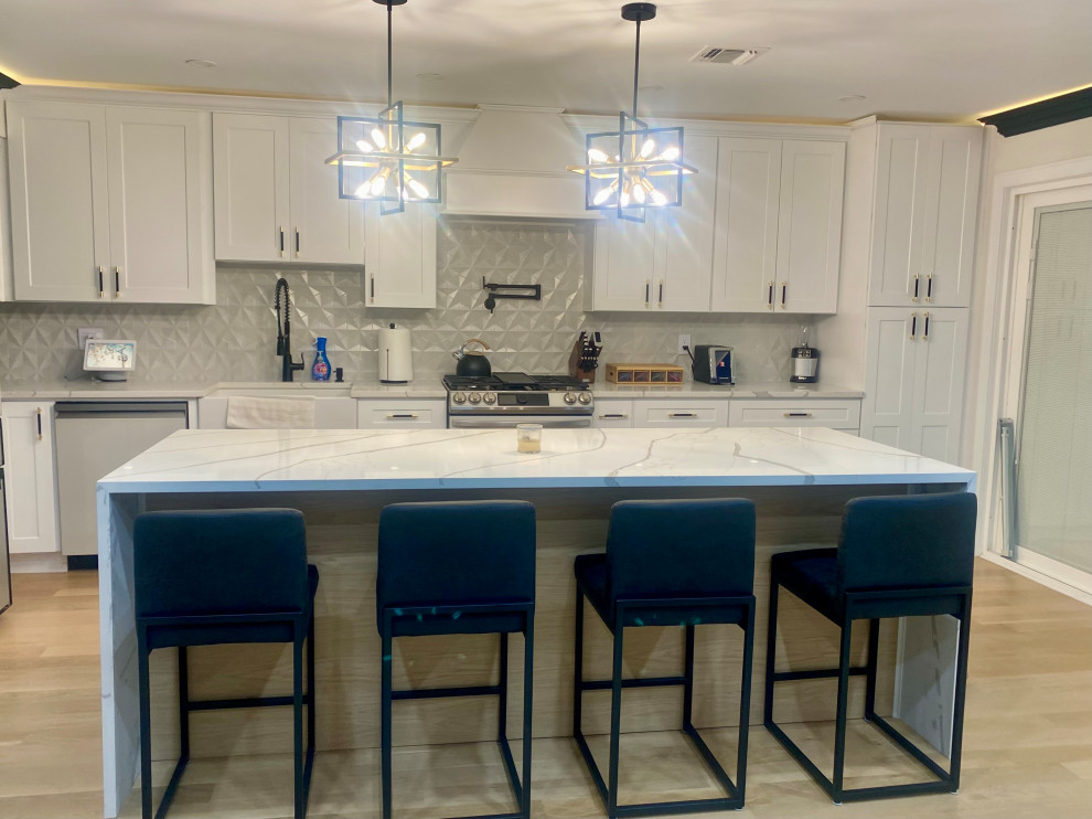 Photo of a large contemporary open plan kitchen in New York with a farmhouse sink, shaker cabinets, white cabinets, quartz benchtops, white splashback, porcelain splashback, stainless steel appliances, light hardwood floors, with island and white benchtop.