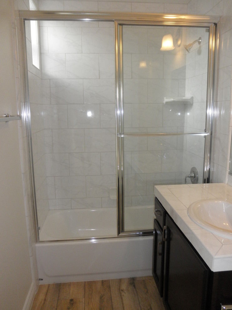 Inspiration for a transitional master beige tile and ceramic tile medium tone wood floor alcove bathtub remodel in Orange County with a drop-in sink, raised-panel cabinets, dark wood cabinets, tile countertops, a two-piece toilet and white walls