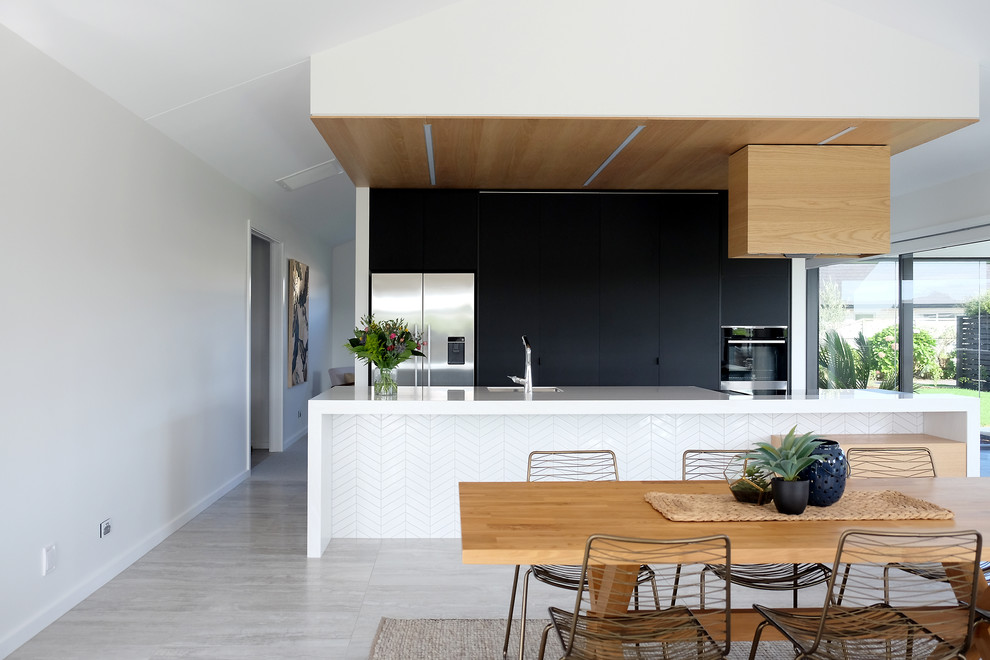 Photo of a mid-sized midcentury galley open plan kitchen in Other with a single-bowl sink, flat-panel cabinets, black cabinets, solid surface benchtops, white splashback, ceramic splashback, stainless steel appliances, ceramic floors, with island, grey floor and white benchtop.