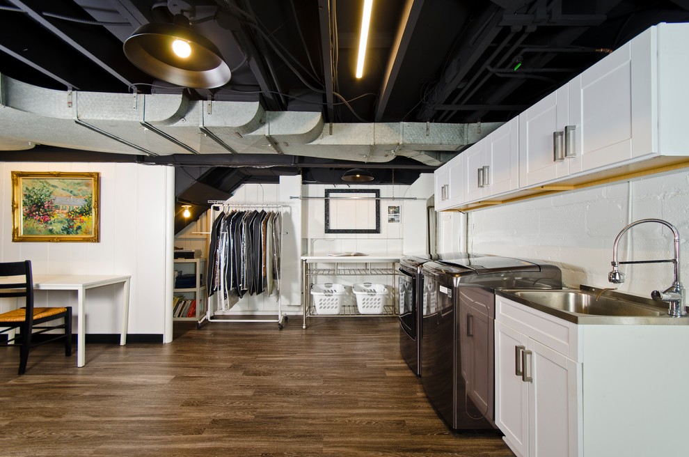 This is an example of a mid-sized industrial utility room in DC Metro with an integrated sink, shaker cabinets, white cabinets, white walls, laminate floors, a side-by-side washer and dryer and brown floor.